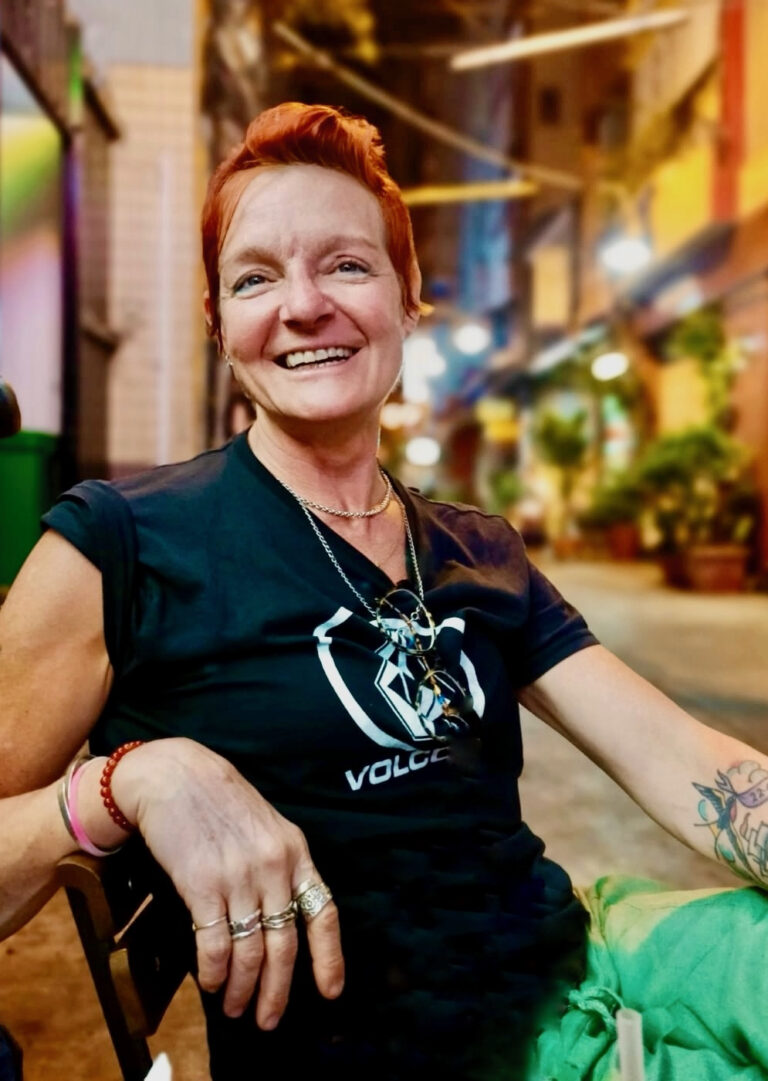 Bil relaxing in a street lit with yellow light, with red hair, black top and green trousers.