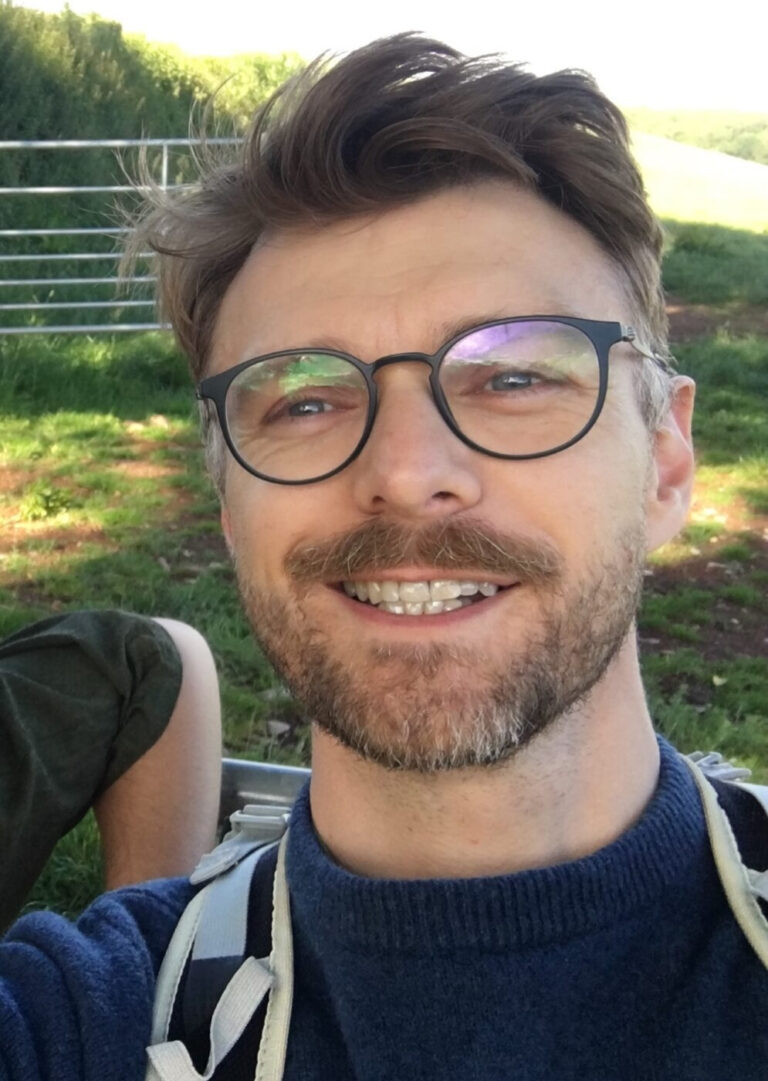 Jacob on a walk in the sunshine, with blond hair, blue glasses, a blue jumper and grey backpack straps.